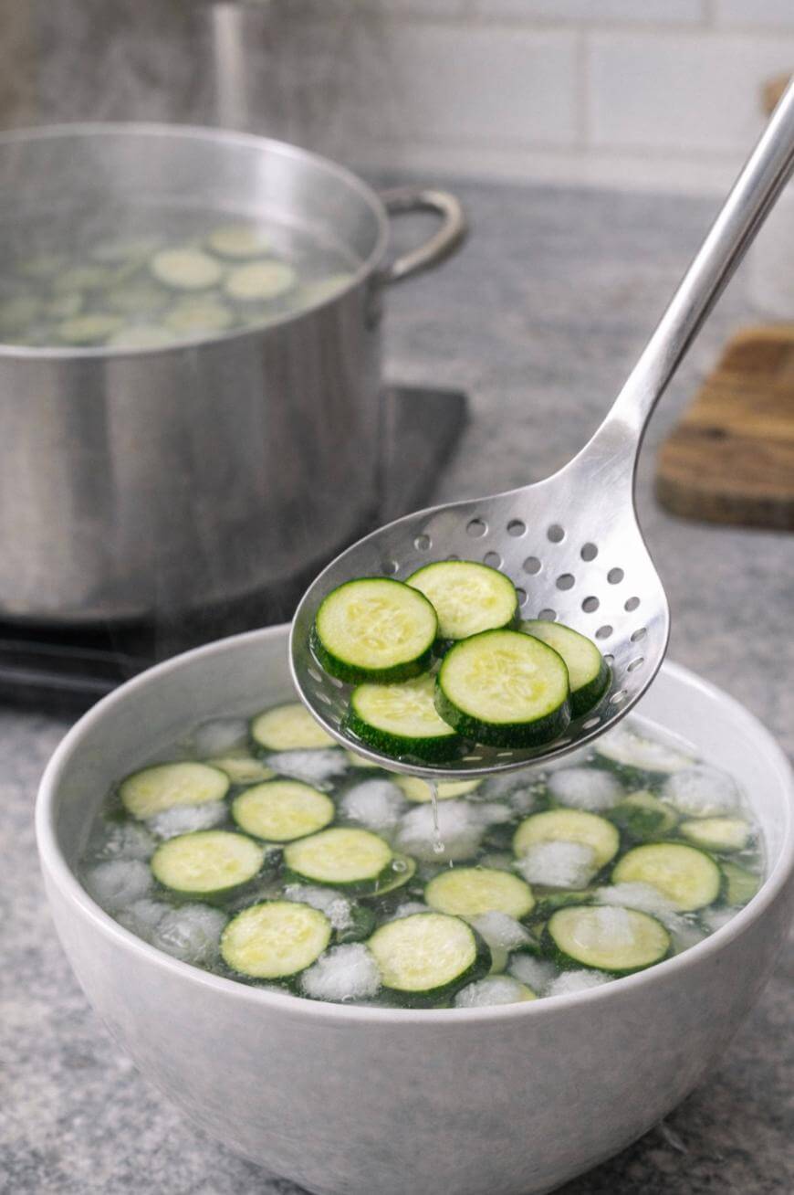 A slotted spoon holds sliced zucchini above a bowl of ice water, with more zucchini and ice cubes floating inside. In the background, a pot of zucchini slices boils on the stove.