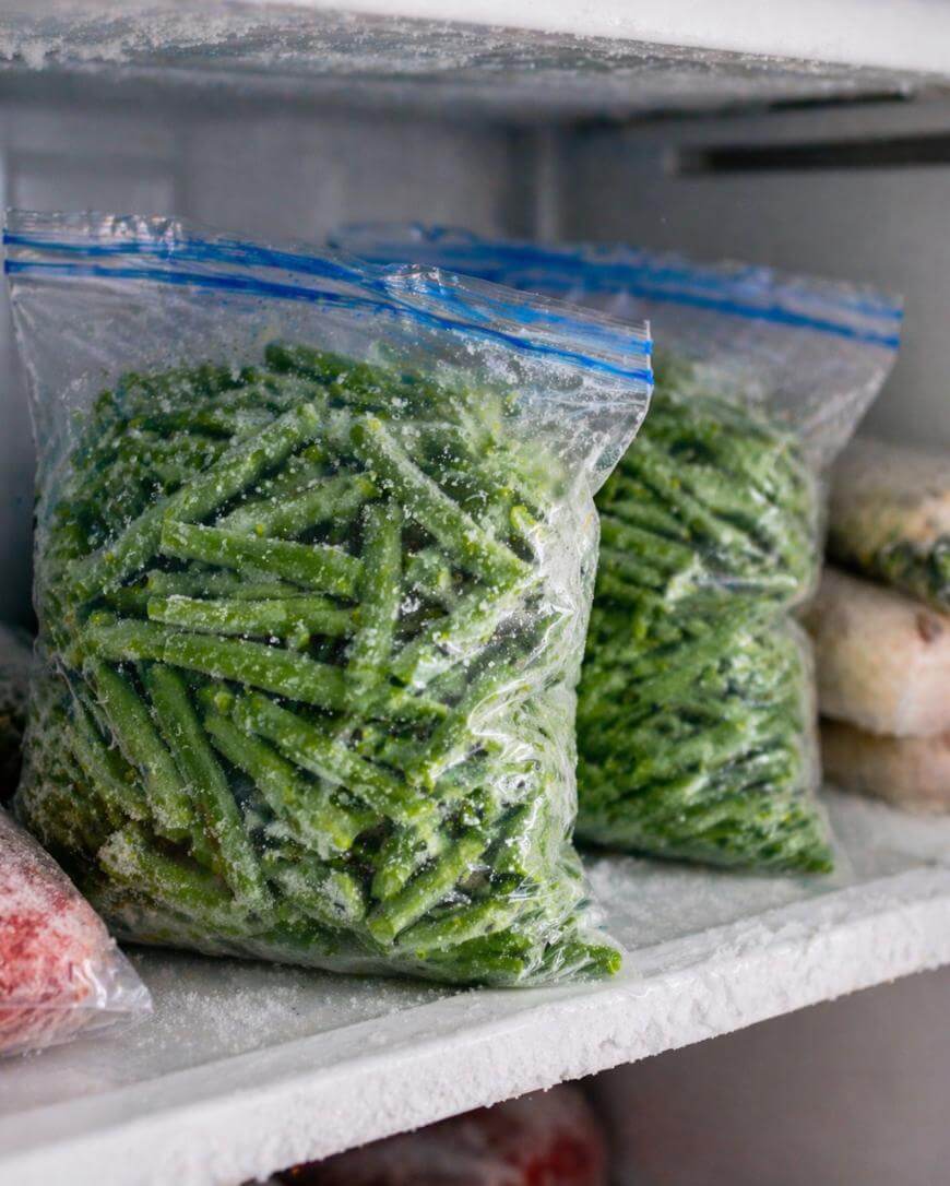 Two freezer bags filled with frozen green beans stand upright on a frosty freezer shelf, surrounded by other frozen food packages.