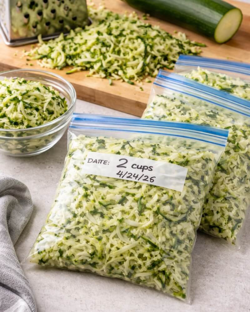 Two labeled freezer bags filled with shredded zucchini sit on a counter next to a glass bowl of more shredded zucchini, a grater, a halved zucchini, and a wooden cutting board in the background.