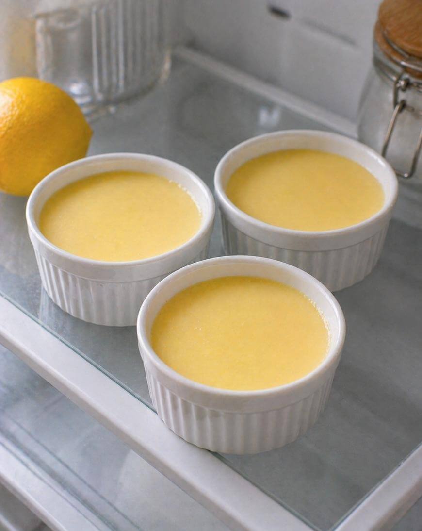 Three white ramekins filled with smooth yellow custard are placed on a glass shelf. A lemon and a glass jar are nearby, suggesting a lemon-flavored dessert.