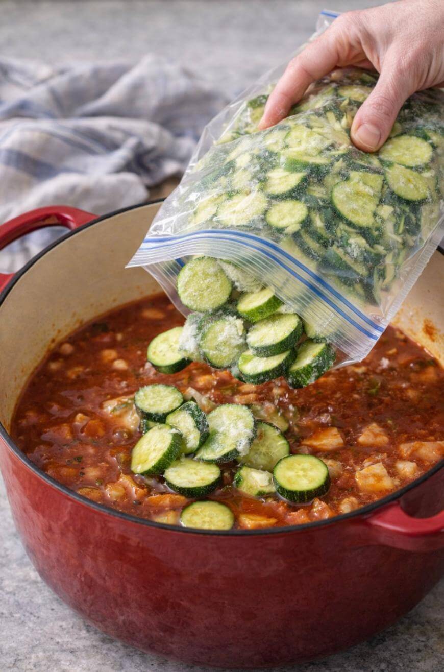 A hand pours frozen sliced zucchini from a plastic bag into a red pot filled with a chunky tomato-based stew. A cloth napkin is in the background on a gray surface.