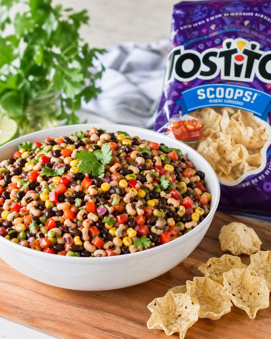 A large white bowl filled with colorful cowboy caviar salad, featuring black beans, black-eyed peas, corn, peppers, and cilantro. Nearby are tortilla chips and a bag of Tostitos Scoops on a wooden board.