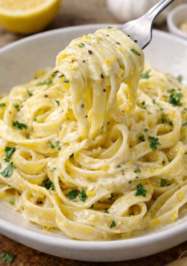 A bowl of creamy fettuccine pasta garnished with chopped parsley and black pepper. A fork is lifting a portion of the pasta, showing the creamy sauce coating the noodles.