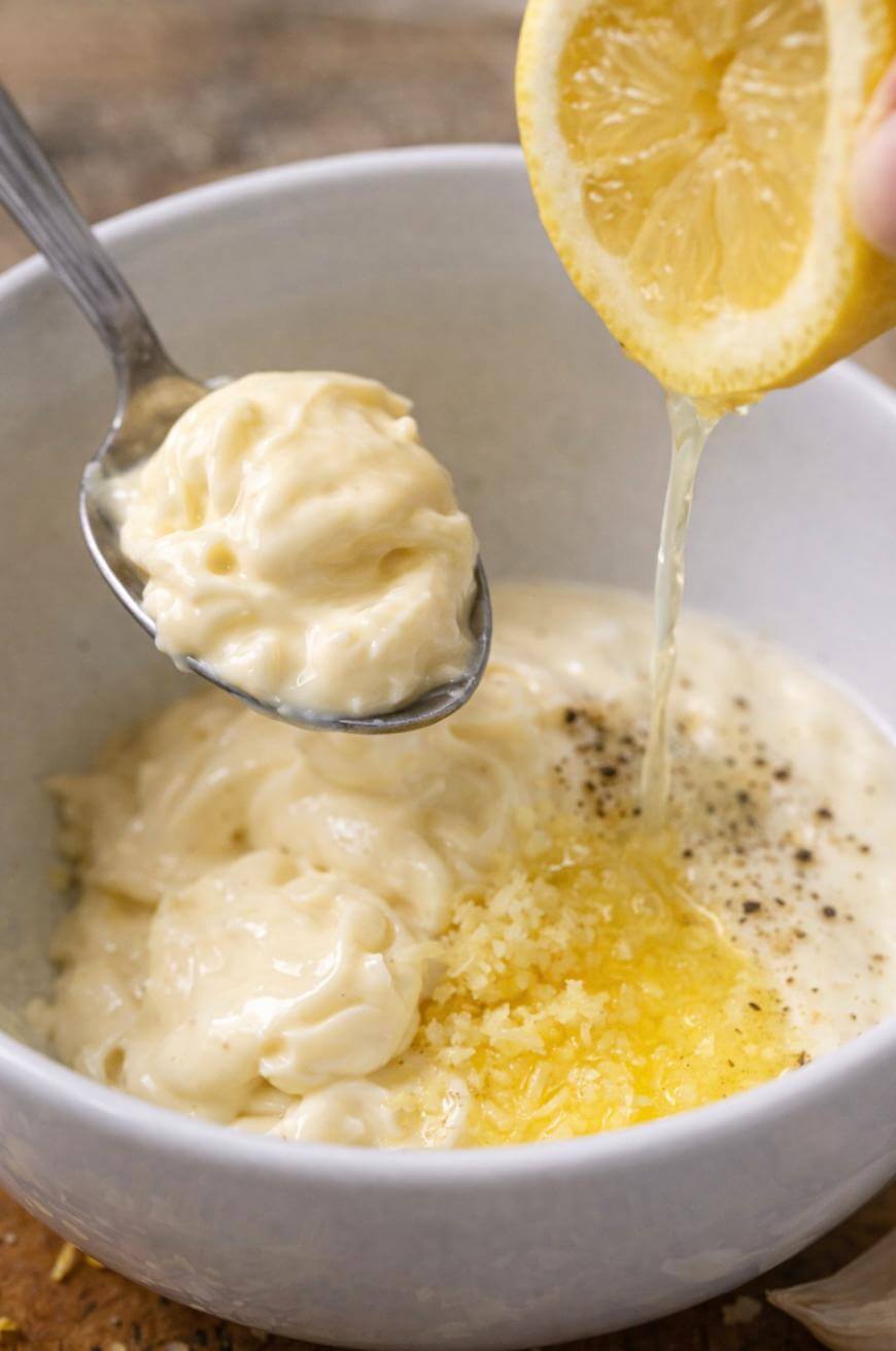 A spoonful of creamy sauce is held over a bowl while fresh lemon juice is being squeezed in. The bowl also contains grated garlic, ground black pepper, and more creamy sauce.