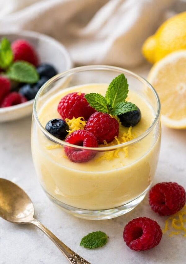 A glass of creamy lemon mousse topped with raspberries, blueberries, lemon zest, and a mint sprig. A gold spoon and more berries are nearby, with lemons and a bowl of berries in the background.