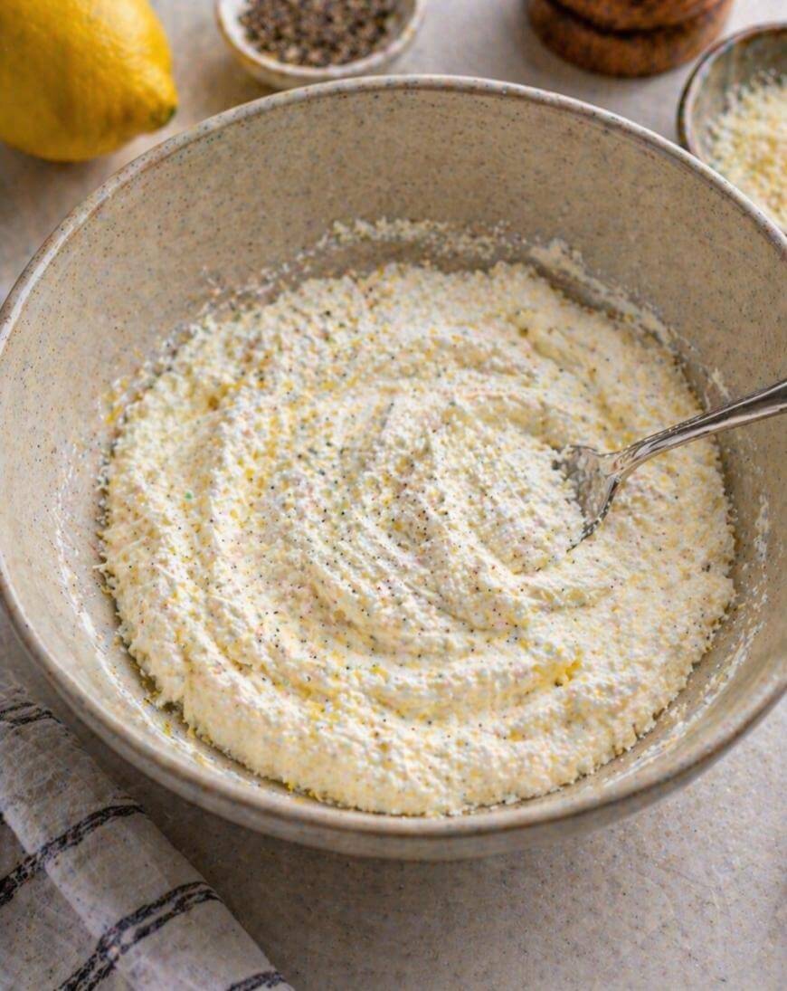 A ceramic bowl filled with a creamy ricotta cheese mixture sits on a countertop, with a metal spoon inside. A lemon, a bowl of pepper, and a striped towel are visible nearby.