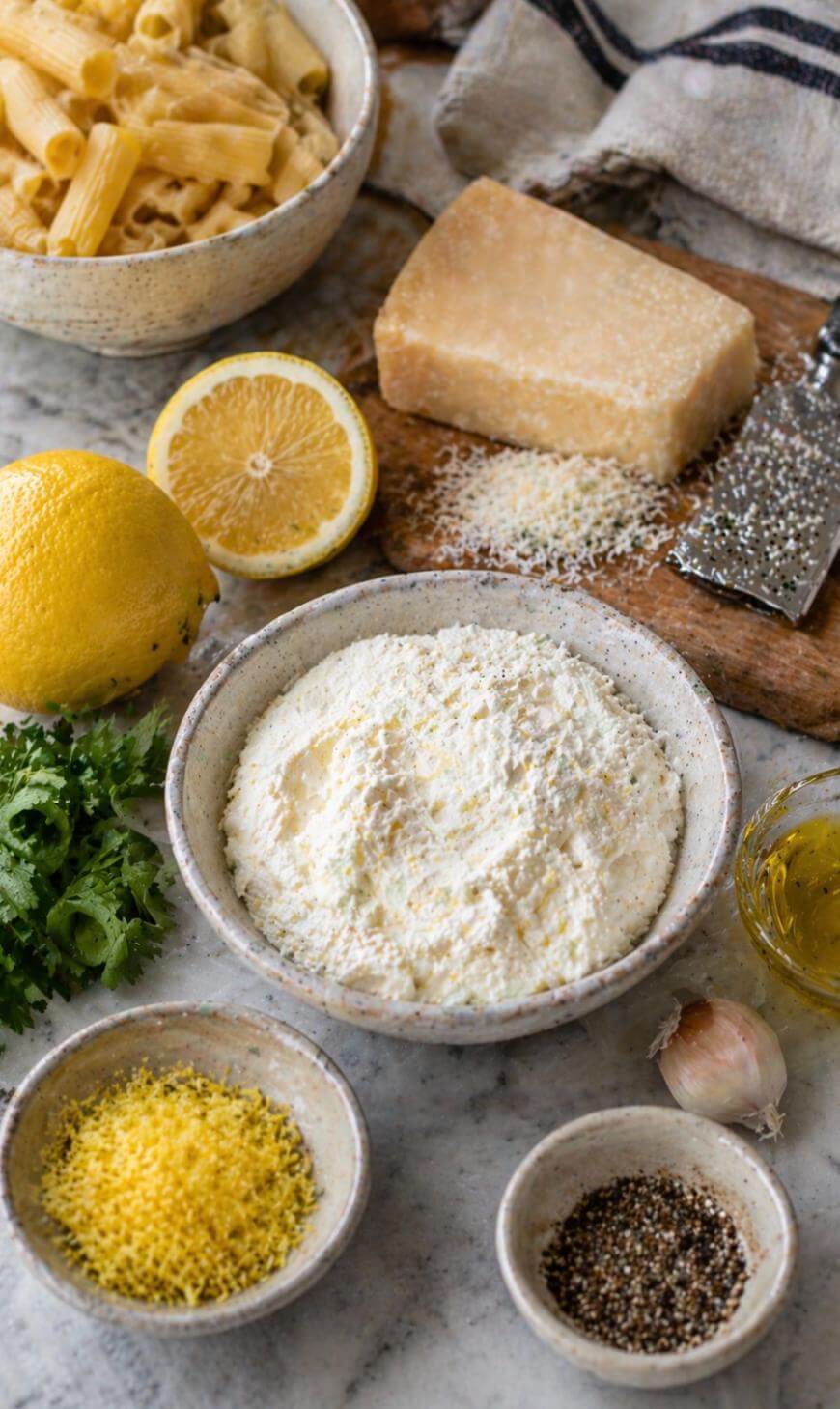 A bowl of ricotta cheese surrounded by cooked pasta, a block of Parmesan, grated cheese, lemon, lemon zest, parsley, black pepper, garlic, and olive oil on a marble countertop.