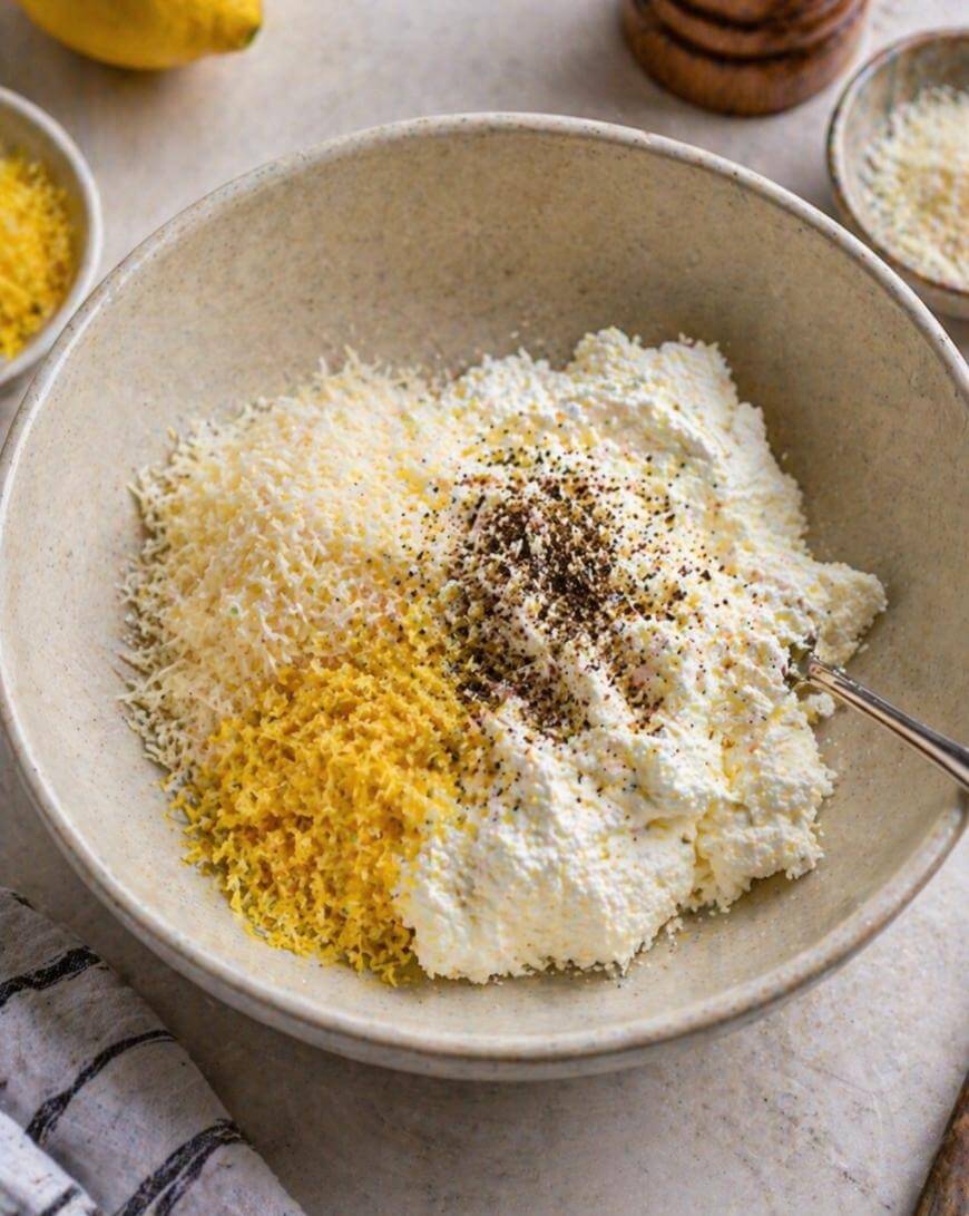 A bowl contains ricotta cheese, grated parmesan, lemon zest, and cracked black pepper, with a spoon resting inside. Other ingredients and a striped towel are nearby.