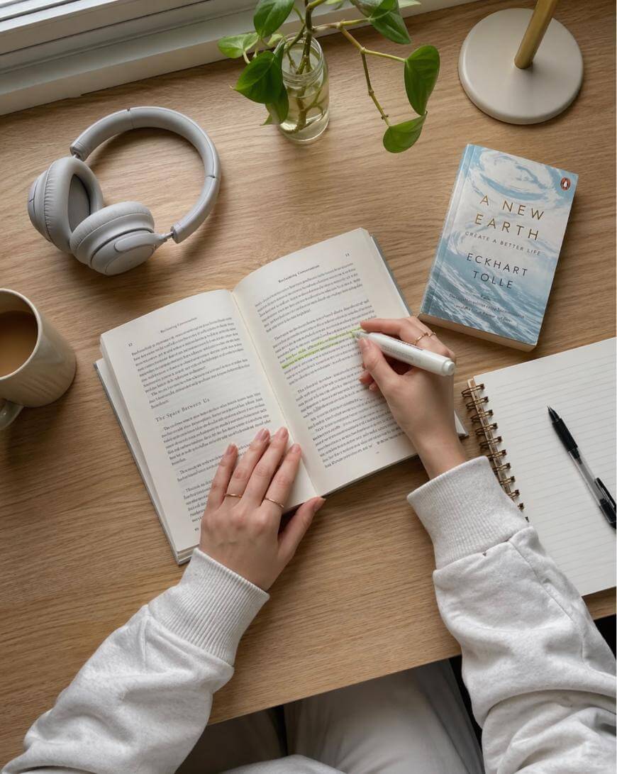 A person in a white sweater highlights text in an open book on a wooden desk with a cup of coffee, headphones, a plant, a notebook with a pen, and a copy of A New Earth by Eckhart Tolle.