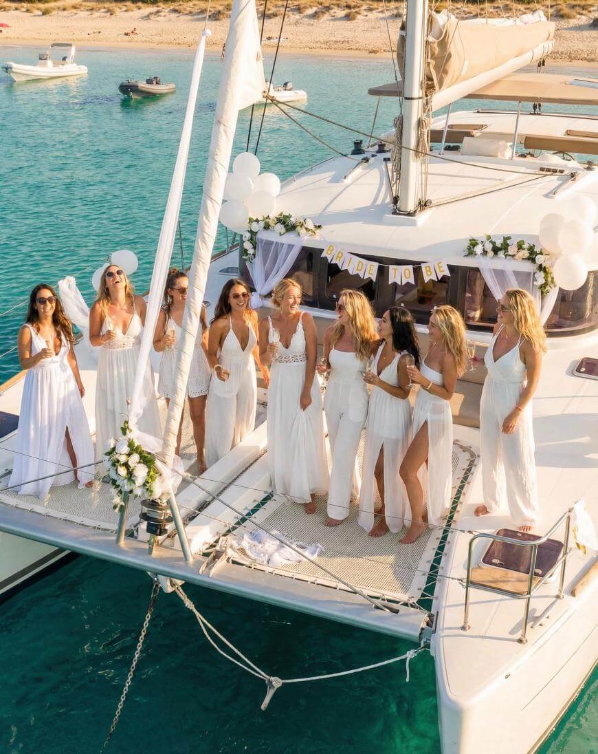 A group of women in white dresses stands on the deck of a boat decorated with flowers and balloons, with clear blue water and a sandy shore in the background, celebrating a bride-to-be.