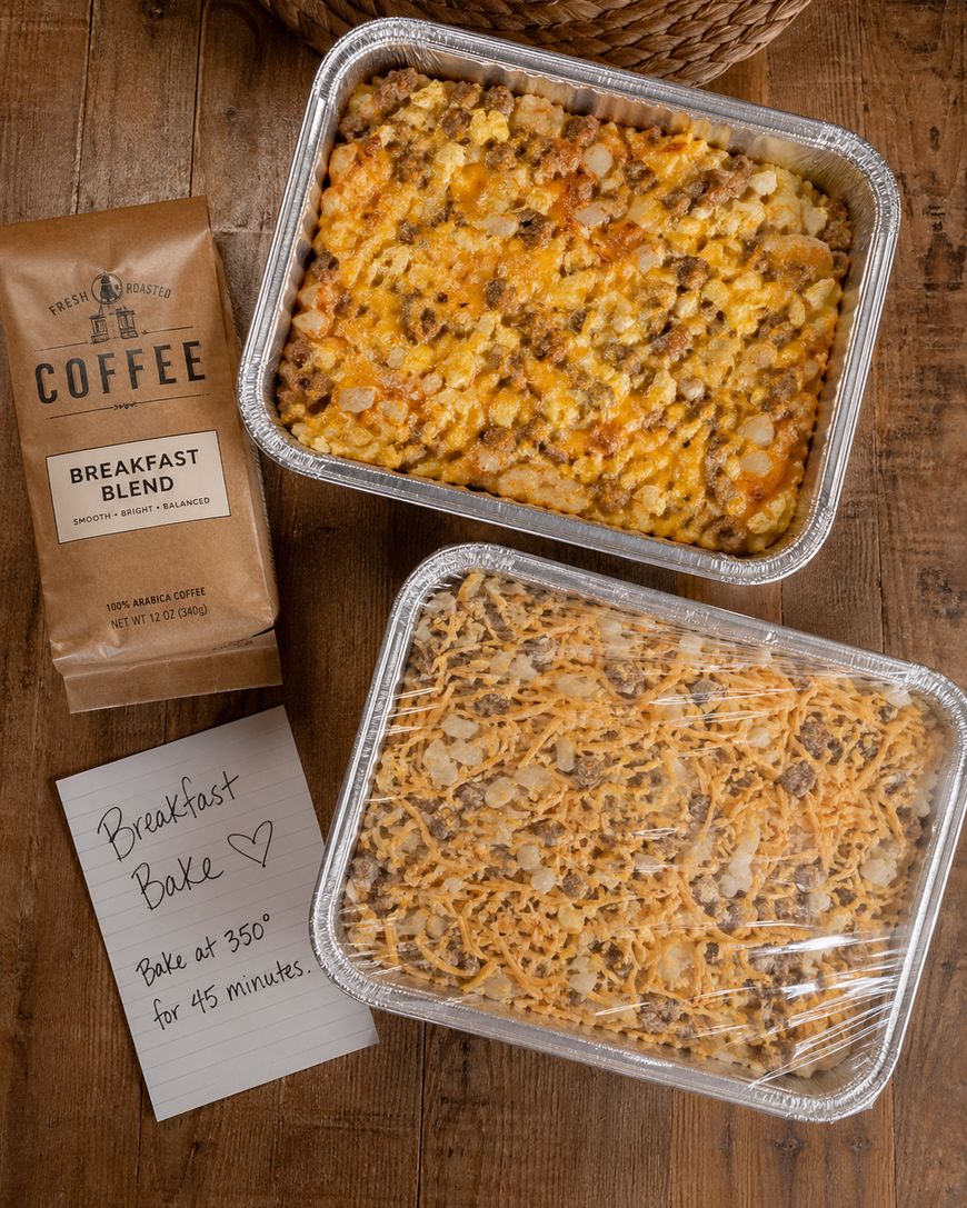 Two aluminum trays of breakfast casserole topped with cheese sit on a wooden table. One tray is covered in plastic wrap, next to a bag of coffee and a handwritten note that says, Breakfast Bake, Bake at 350° for 45 minutes.