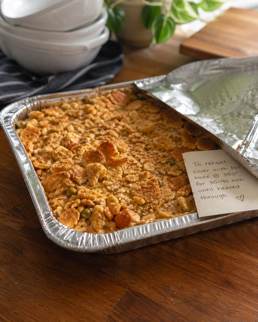 A foil baking tray filled with a golden, crumb-topped casserole sits on a wooden counter, partly covered with foil. A handwritten note gives reheating instructions. Stacked white bowls and a plant are in the background.