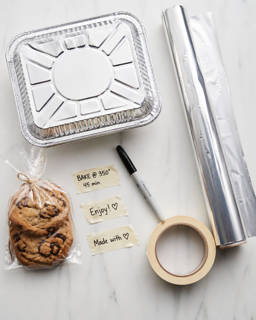 A disposable foil pan with lid, roll of aluminum foil, round masking tape, black marker, three chocolate chip cookies in a tied plastic bag, and three handwritten labels on a white marble surface.