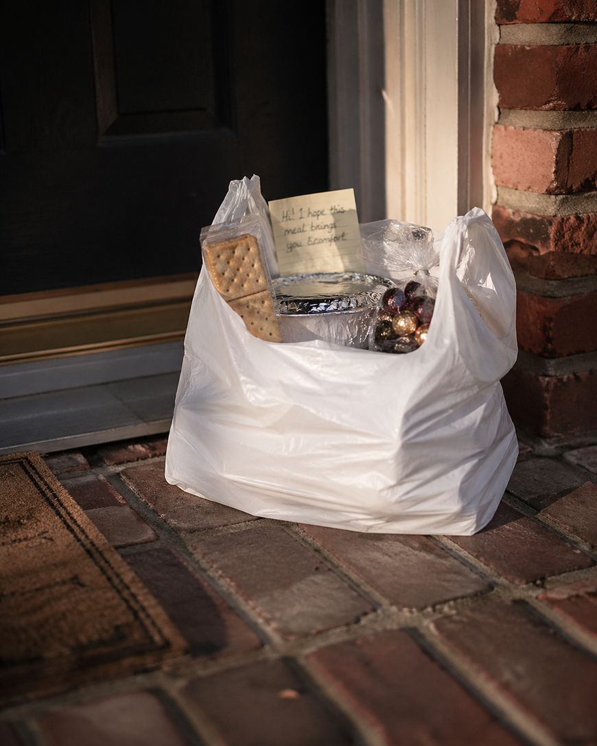 A white plastic bag with food containers, crackers, and a handwritten note sits on a brick doorstep next to a welcome mat and a black door.