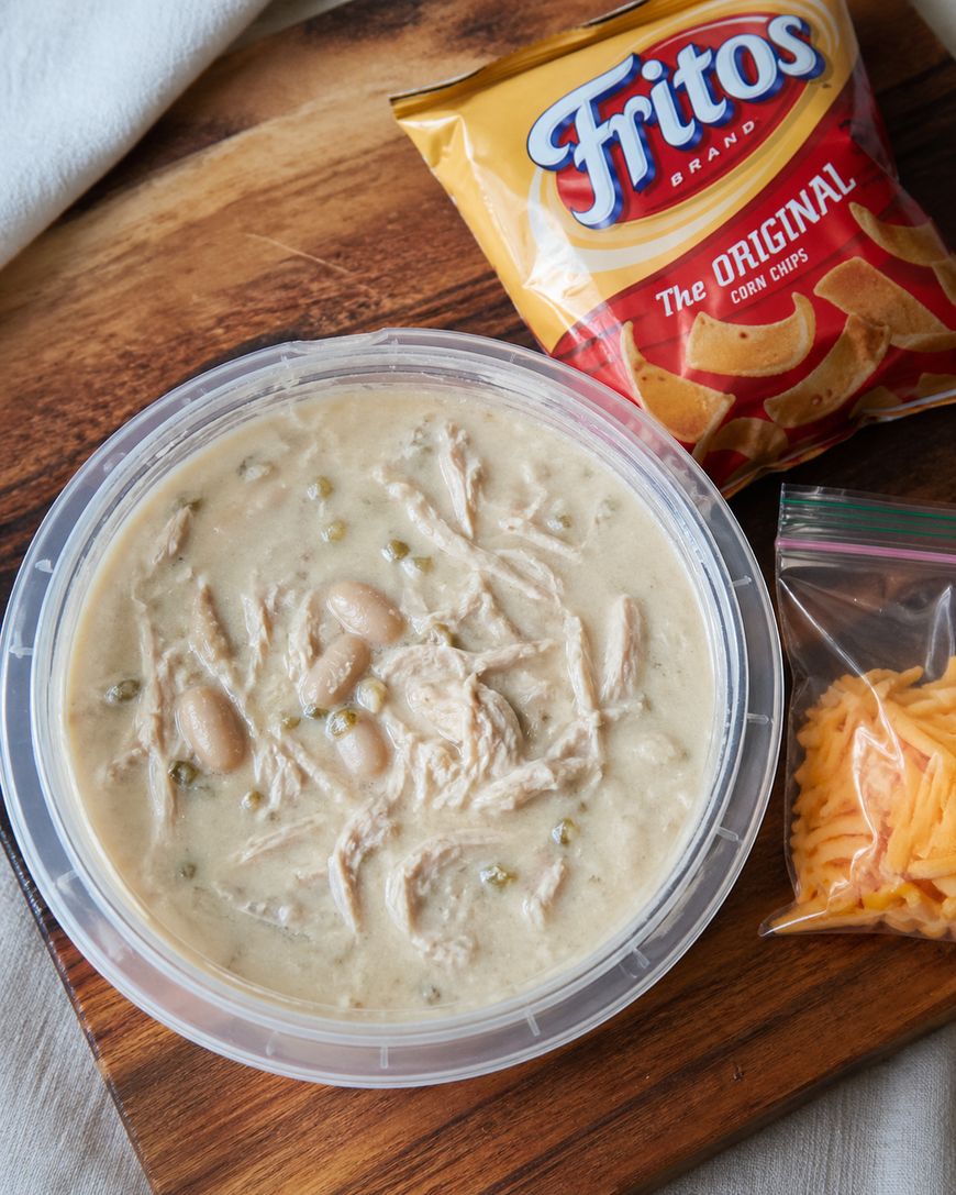 A container of white chicken chili with beans sits on a wooden surface, next to a bag of Fritos corn chips and a small ziplock bag of shredded cheddar cheese.