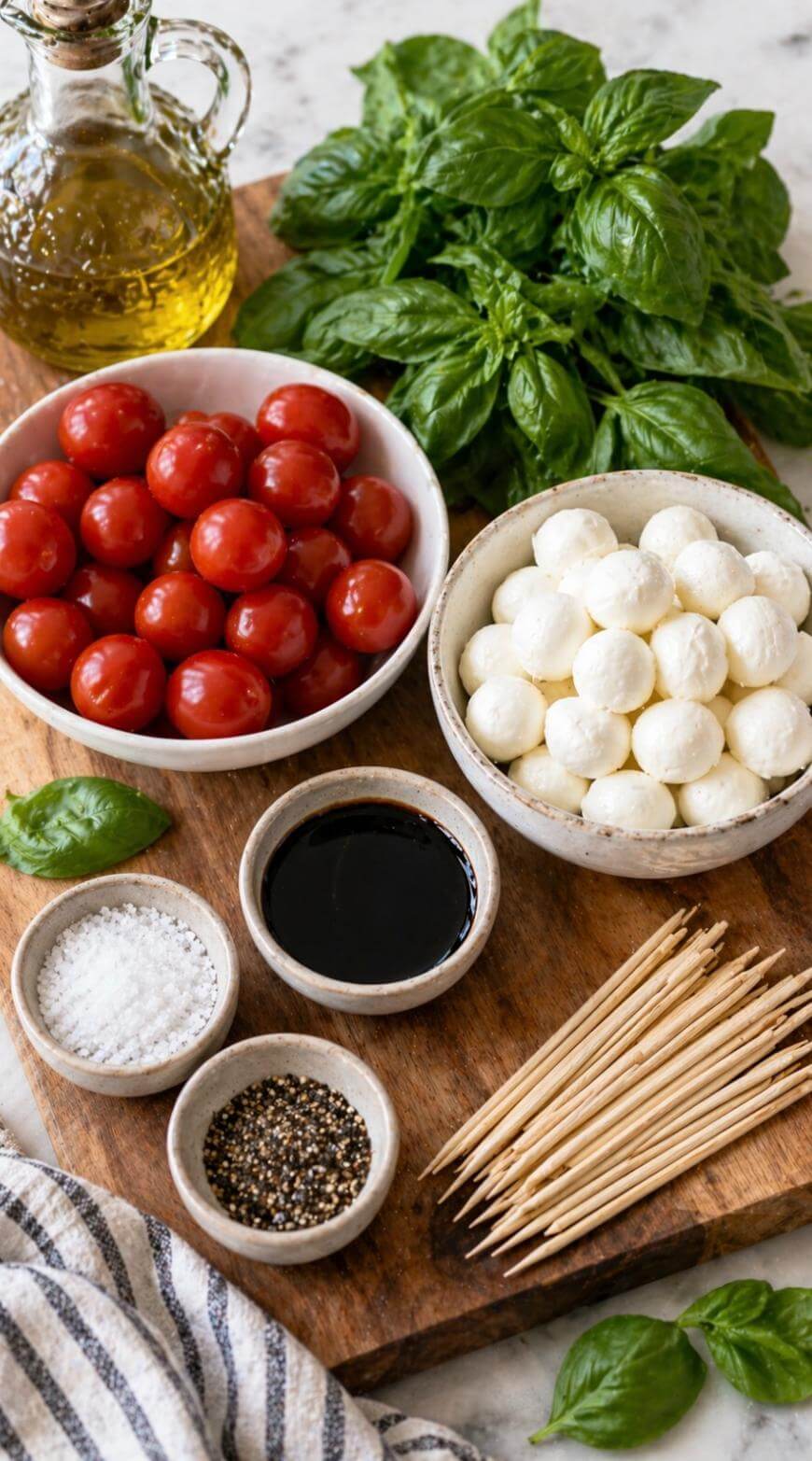 A wooden board with bowls of cherry tomatoes, mozzarella balls, basil leaves, olive oil, balsamic vinegar, salt, pepper, and wooden toothpicks, ready for making caprese appetizers.