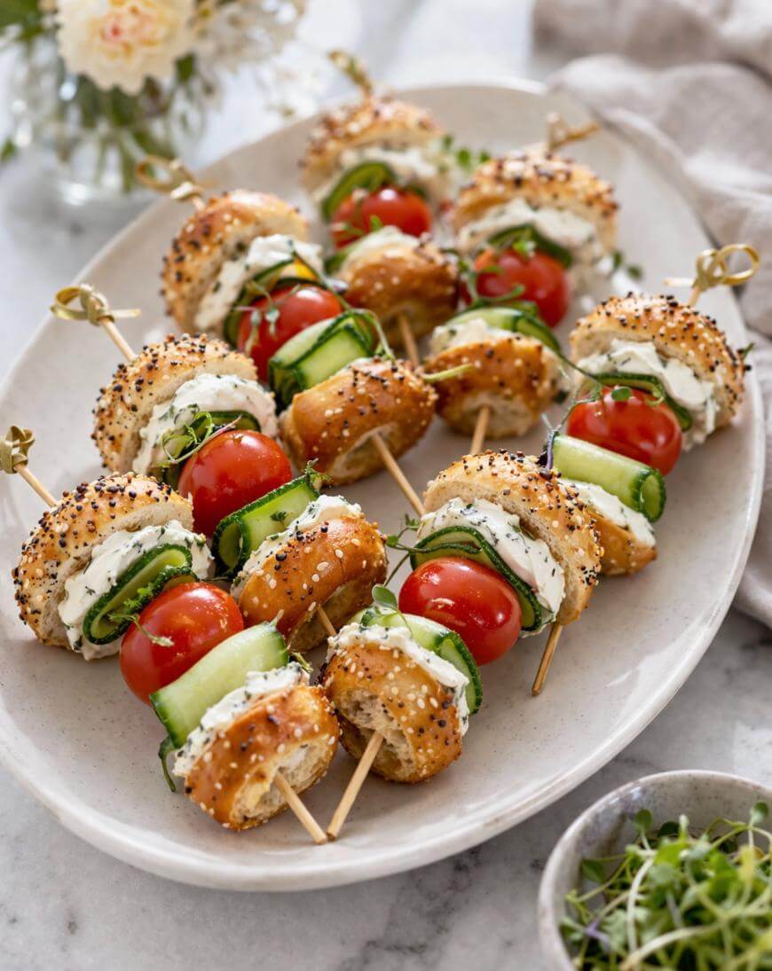 Skewers with mini bagel halves, cream cheese, cucumber slices, and cherry tomatoes, arranged on a white oval platter. Microgreens are sprinkled on top, and a flower vase and napkin are in the background.