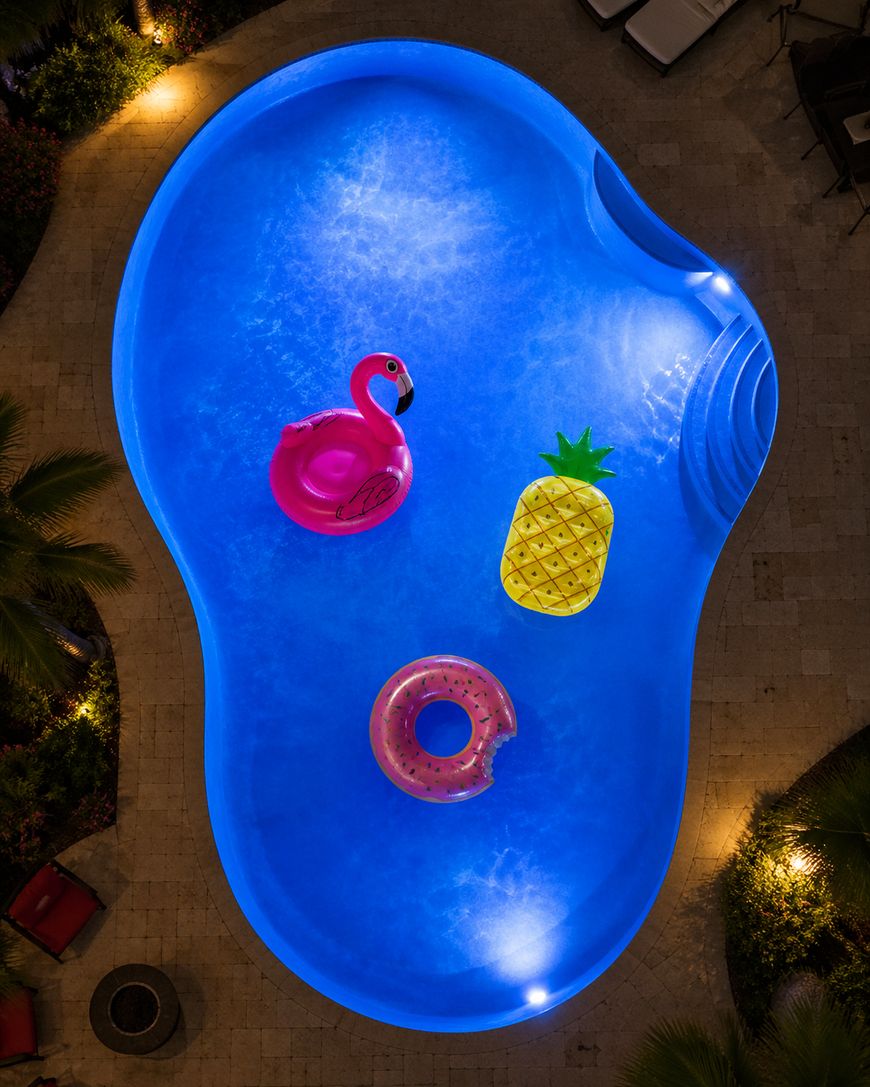 Aerial view of a glowing blue swimming pool at night with three pool floats: a pink flamingo, a yellow pineapple, and a pink frosted donut. The pool is surrounded by plants and lounge chairs.