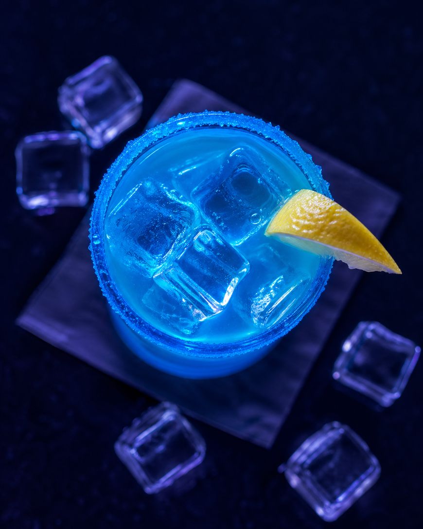 A top-down view of a bright blue cocktail with ice cubes, a sugared rim, and a lemon wedge garnish, surrounded by a few ice cubes on a dark surface.