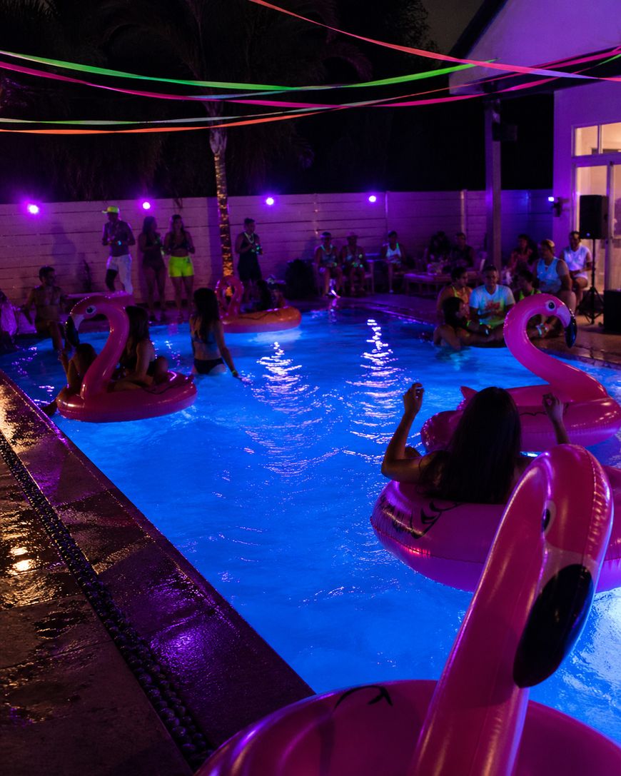 People enjoying a nighttime pool party with glowing lights, colorful neon decorations, and large pink flamingo floaties in the water. The pool and area are illuminated by purple and blue lighting.