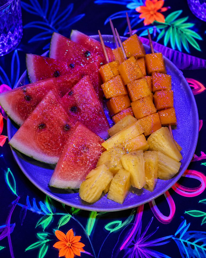 A plate of fresh fruit sits on a colorful, floral-patterned tablecloth. The plate contains watermelon slices, pineapple chunks, and mango cubes on skewers, all sparkling under vibrant lighting.