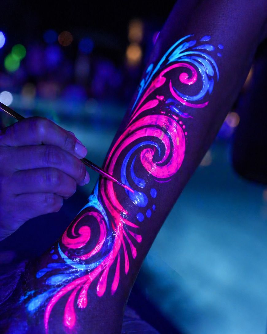 A hand paints vibrant neon pink and blue swirling patterns on a persons leg, glowing under blacklight with a blurred background, possibly near a pool at night.