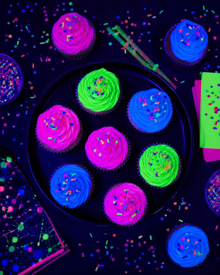 A black plate holds nine neon cupcakes with pink, blue, and green frosting, topped with colorful sprinkles. Neon confetti, napkins, and cups surround the plate on a dark background.
