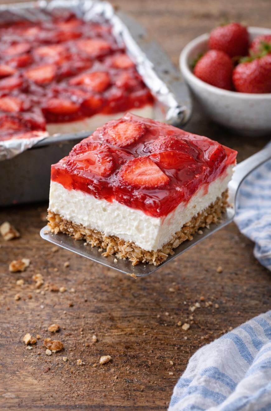 A spatula holds a square piece of layered dessert with a crumbly crust, creamy middle, and strawberry topping; a pan of dessert and a bowl of strawberries sit in the background on a wooden table.