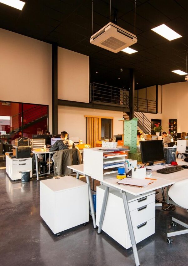 Modern open office space with white desks, computers, and a few people working. The room has high ceilings, bright lighting, large windows, and a mezzanine with metal railings above.