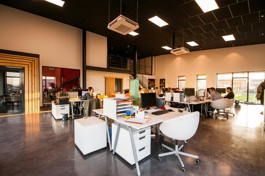 Modern open-plan office with several desks and office chairs. People are working on computers in a bright, spacious room with high ceilings, large windows, and a loft mezzanine. Office equipment and paperwork are visible.