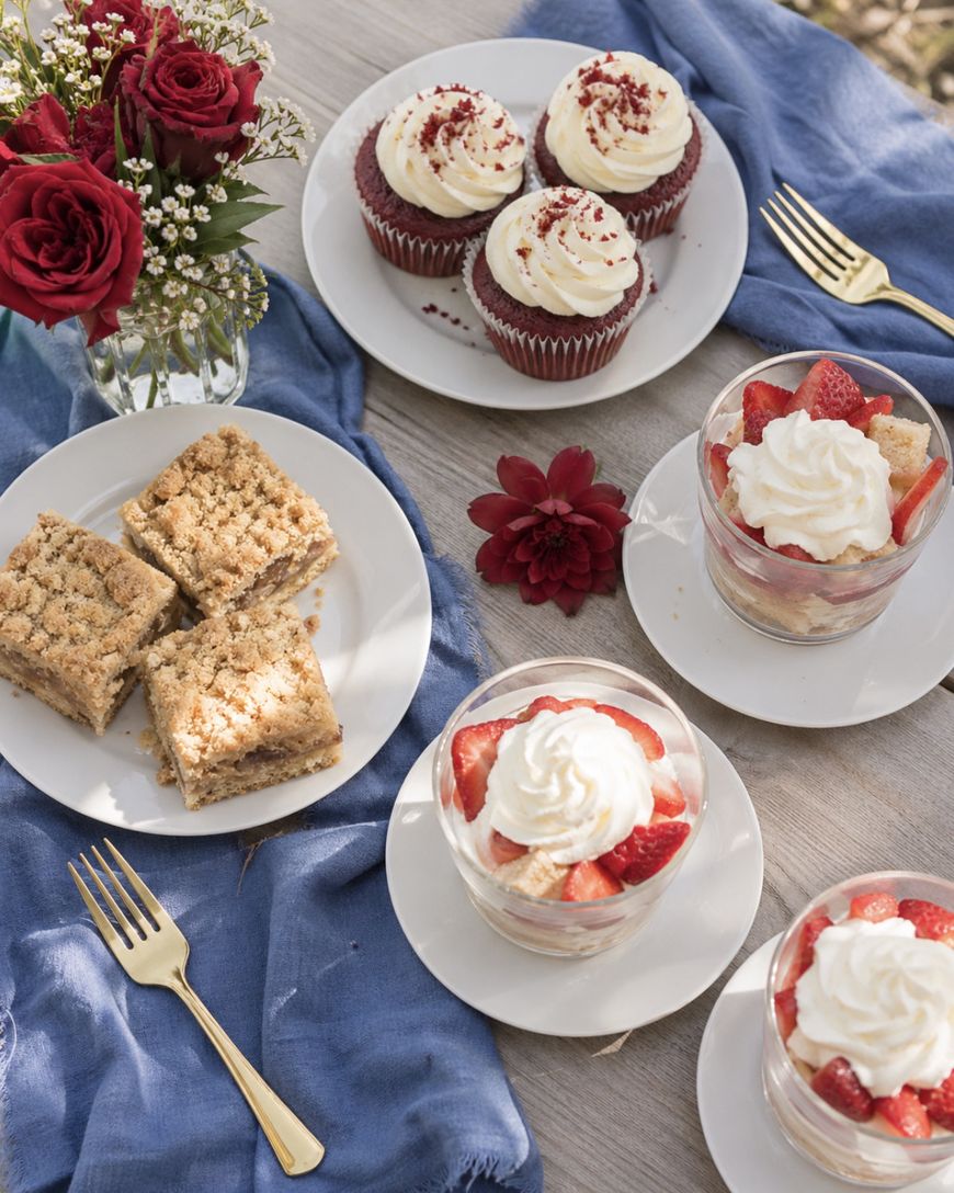 A tabletop with red velvet cupcakes, strawberry shortcake parfaits, crumb cake squares, gold forks, red roses in a vase, and a blue cloth, arranged for a romantic dessert setting outdoors.