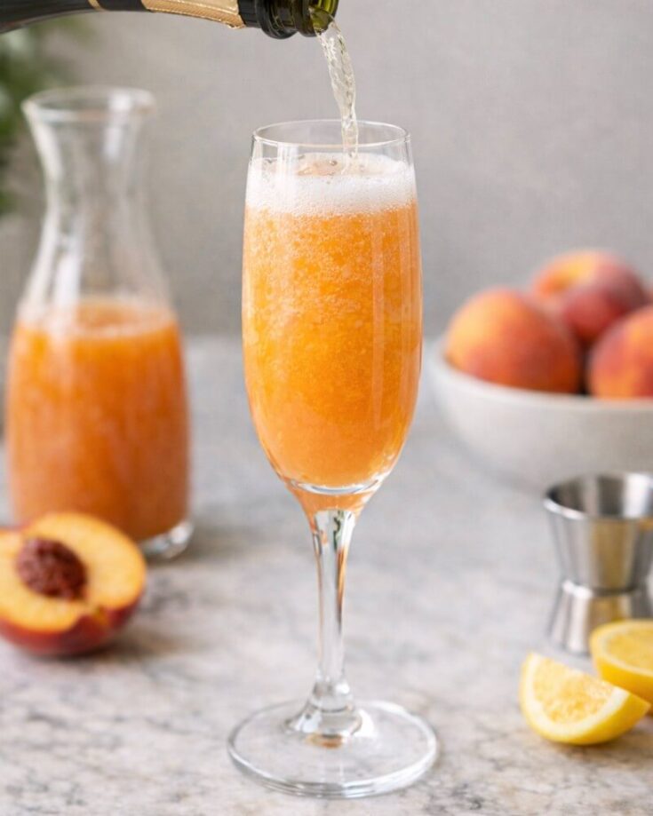 A sparkling drink is being poured into a champagne flute filled with a peach-colored beverage. In the background, there is a carafe of the same drink, a halved peach, lemon wedges, and a bowl of whole peaches.