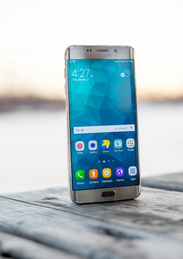 A Samsung smartphone standing upright on a wooden surface outdoors, displaying its home screen with various app icons and the time 4:27, against a blurred background.