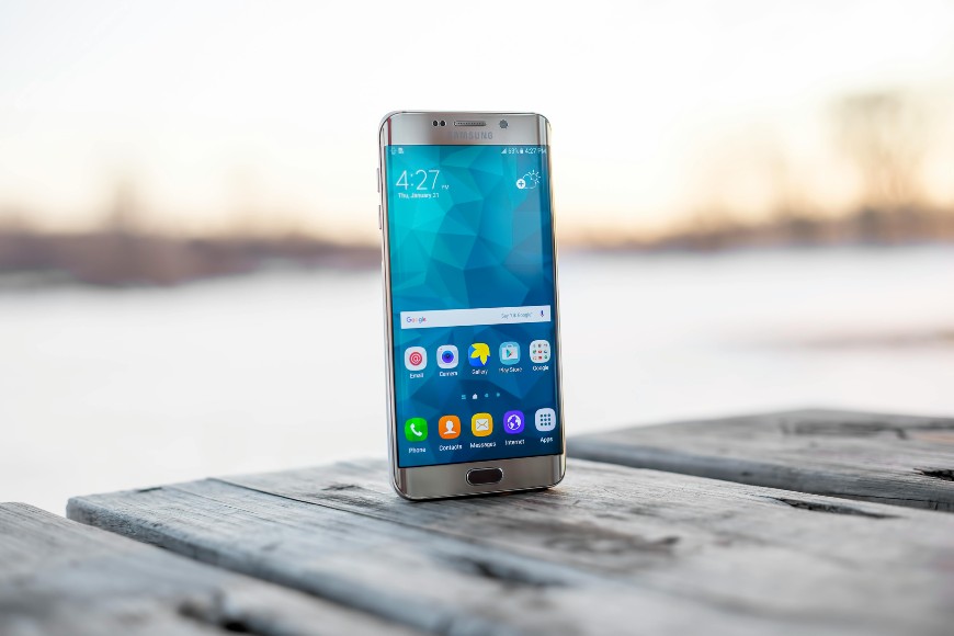 A smartphone displaying its home screen stands upright on a weathered wooden surface outdoors, with a blurred background of trees and a bright sky.