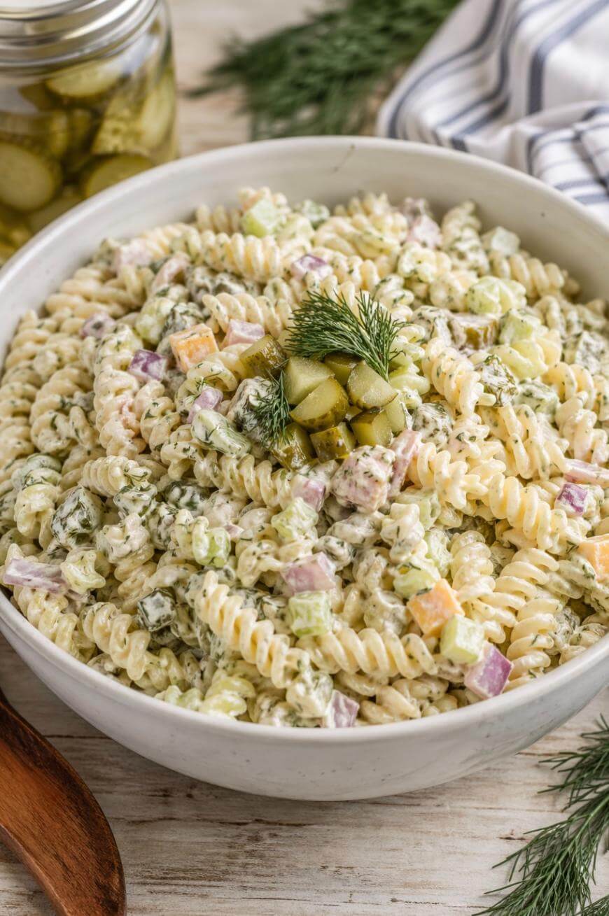 A bowl of creamy rotini pasta salad with diced pickles, red onions, celery, and cheddar cheese, garnished with fresh dill and pickle pieces. A jar of pickles and striped cloth are in the background.