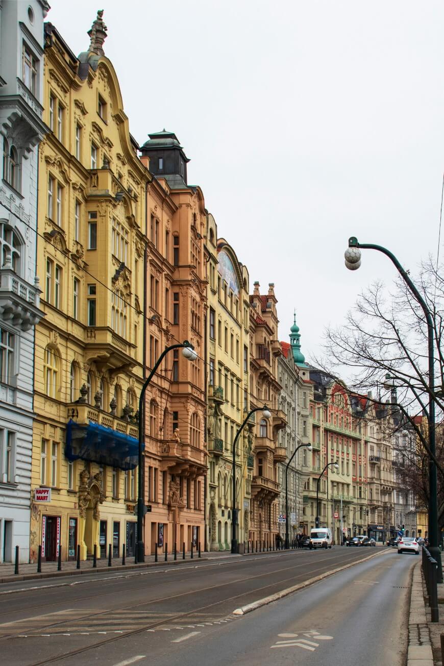 A street lined with colorful historic buildings featuring ornate facades and arched windows, with parked cars along the curb and streetlights overhead on a cloudy day.