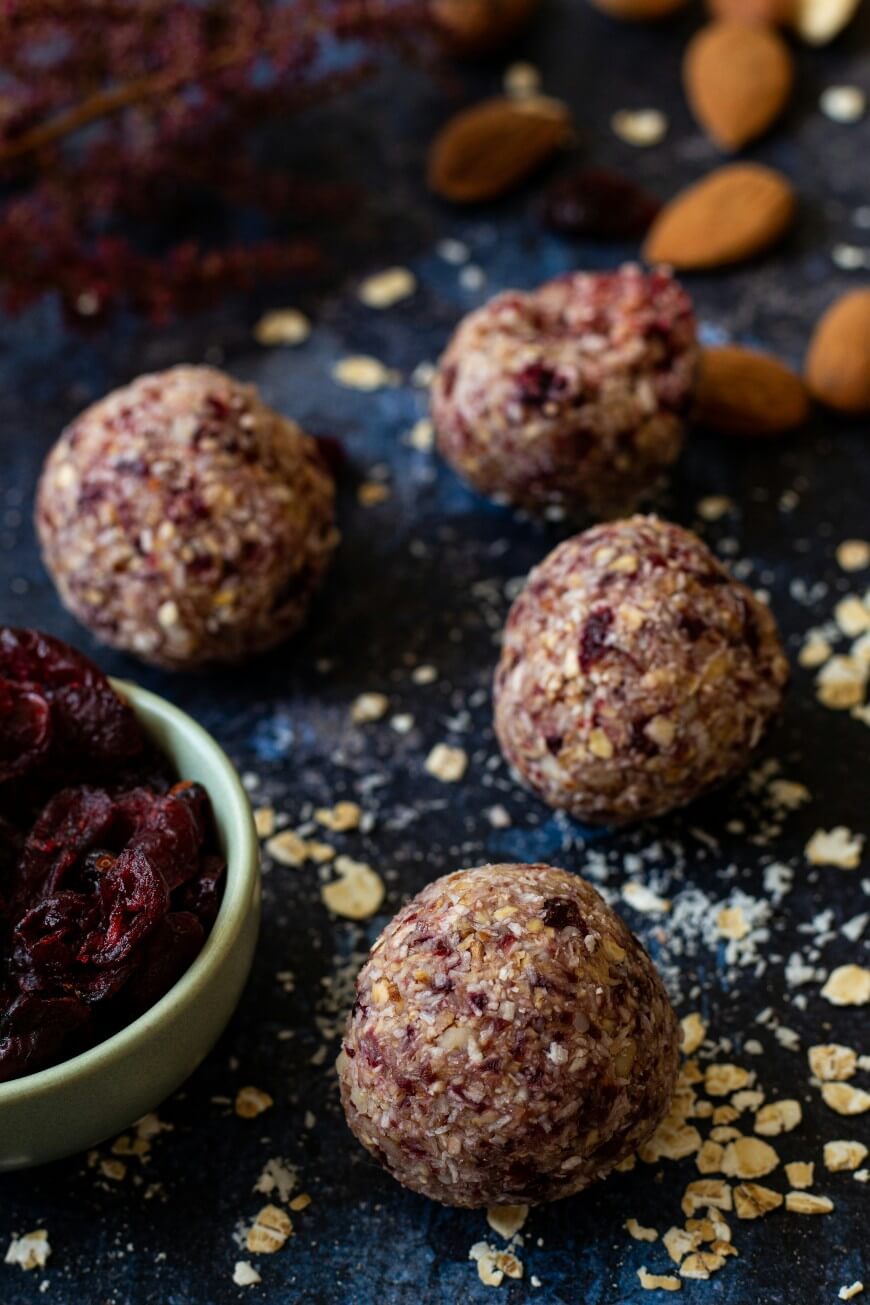 Four homemade energy balls with oats and dried cranberries are arranged on a dark surface, surrounded by scattered oats, whole almonds, and a bowl of dried cranberries.