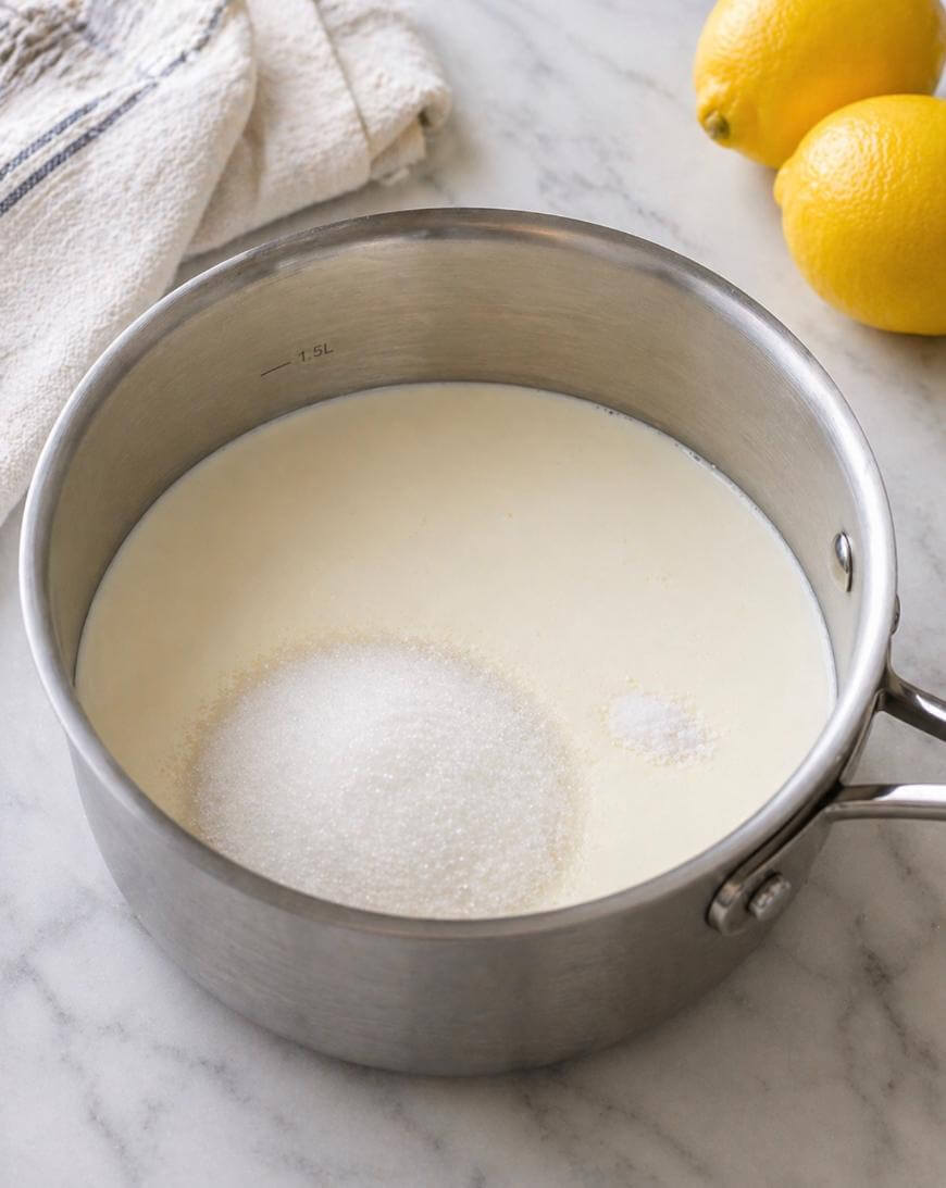 A stainless steel saucepan filled with milk, granulated sugar, and salt sits on a marble countertop. Two lemons and a folded kitchen towel are in the background.