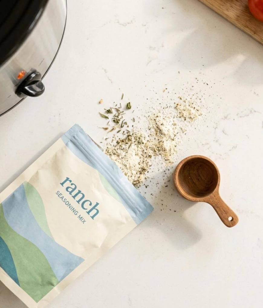 A packet of ranch seasoning mix is open on a white countertop, with some of the mix and herbs spilled next to a small wooden measuring cup. Part of a slow cooker and a wooden board are visible nearby.