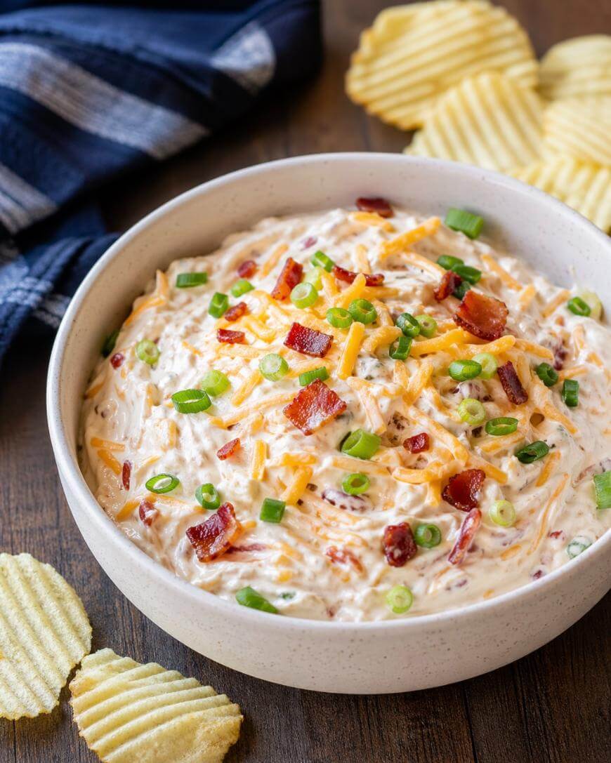 A bowl of creamy dip topped with shredded cheese, bacon bits, and chopped green onions, surrounded by ridged potato chips on a wooden surface. A blue plaid cloth is partially visible in the background.