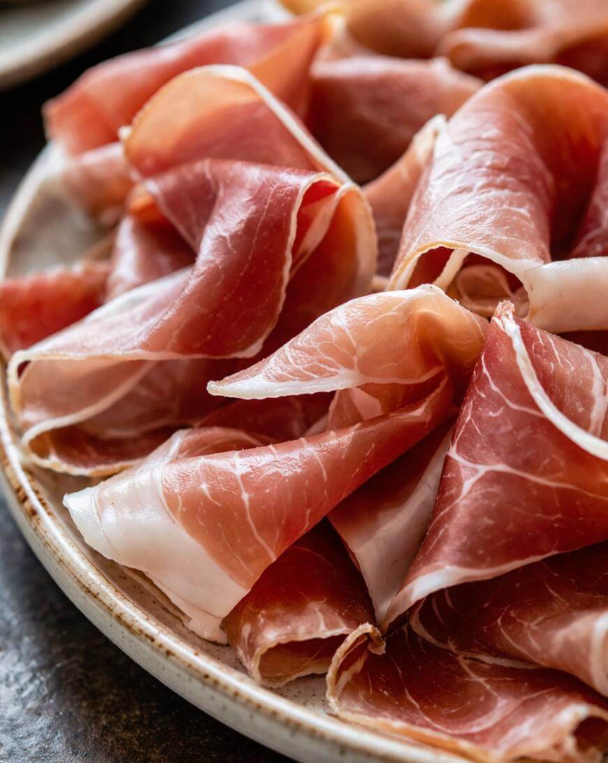 A close-up of thinly sliced prosciutto arranged in delicate folds on a beige ceramic plate.