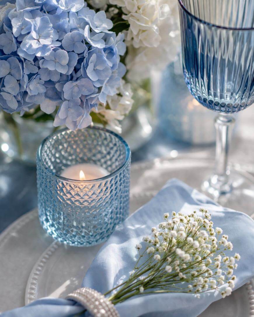A close-up of a table setting with blue hydrangeas, a lit tealight in a blue glass holder, a blue wine glass, and a napkin with a sprig of white babys breath flowers on a glass plate.