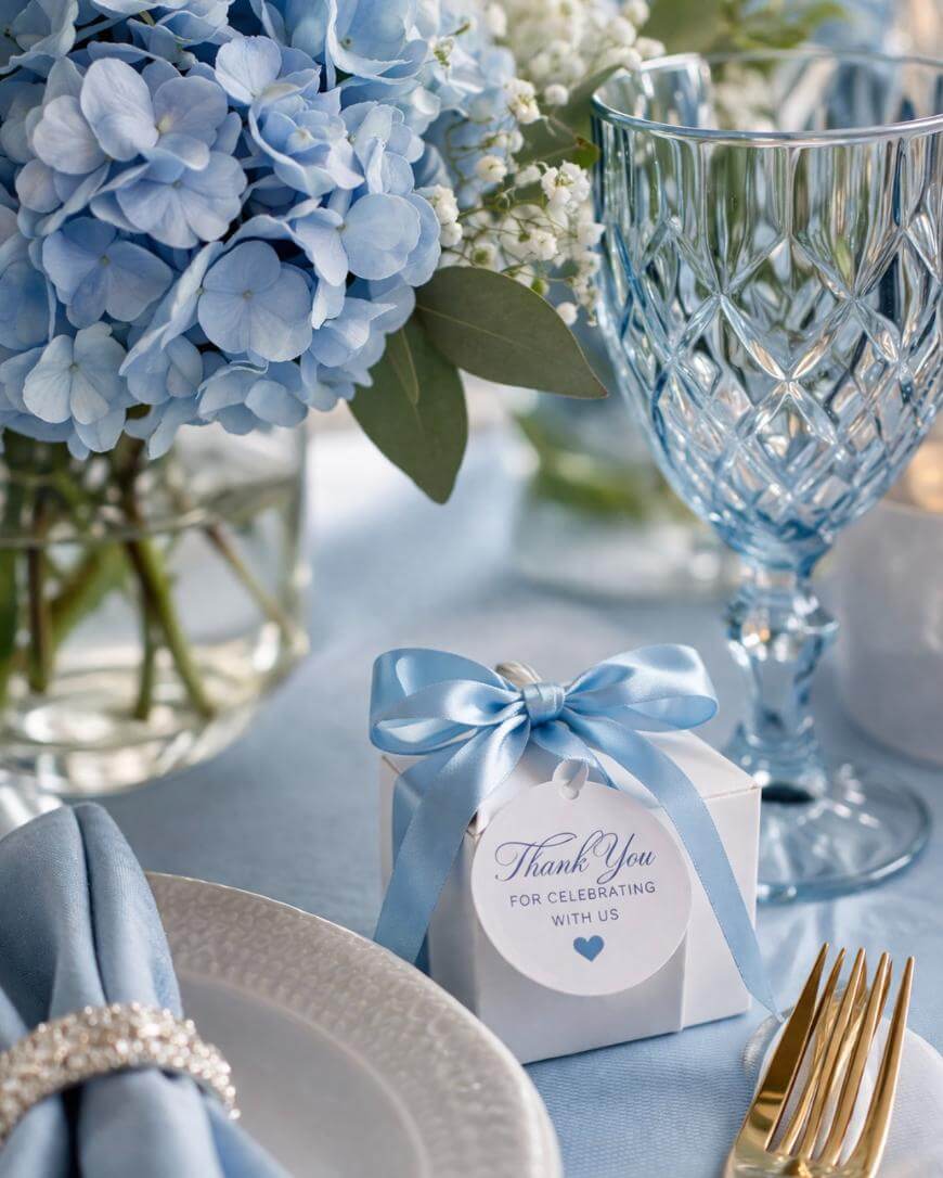 A table setting with blue hydrangeas in a vase, crystal glasses, a white gift box with a blue ribbon that says Thank You for celebrating with us, a white napkin with a jeweled ring, and gold cutlery.