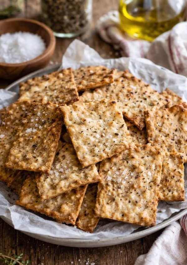 A plate of homemade rectangular crackers sprinkled with coarse salt and herbs, arranged on parchment paper. In the background are jars of salt, pepper, and a bottle of olive oil.