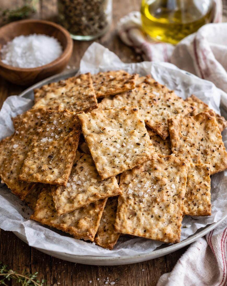 A plate of homemade square crackers topped with sea salt and herbs, arranged on parchment paper. In the background are a glass jar of salt, a bottle of olive oil, and a sprig of fresh thyme.