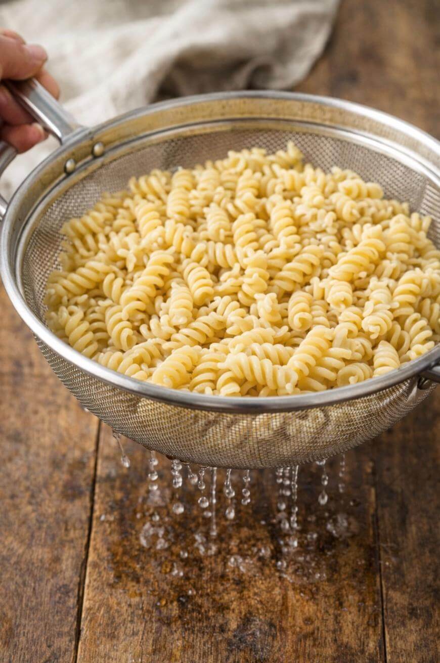 A hand holds a metal colander filled with cooked rotini pasta, with water draining through the mesh over a rustic wooden surface. A beige cloth is visible in the background.