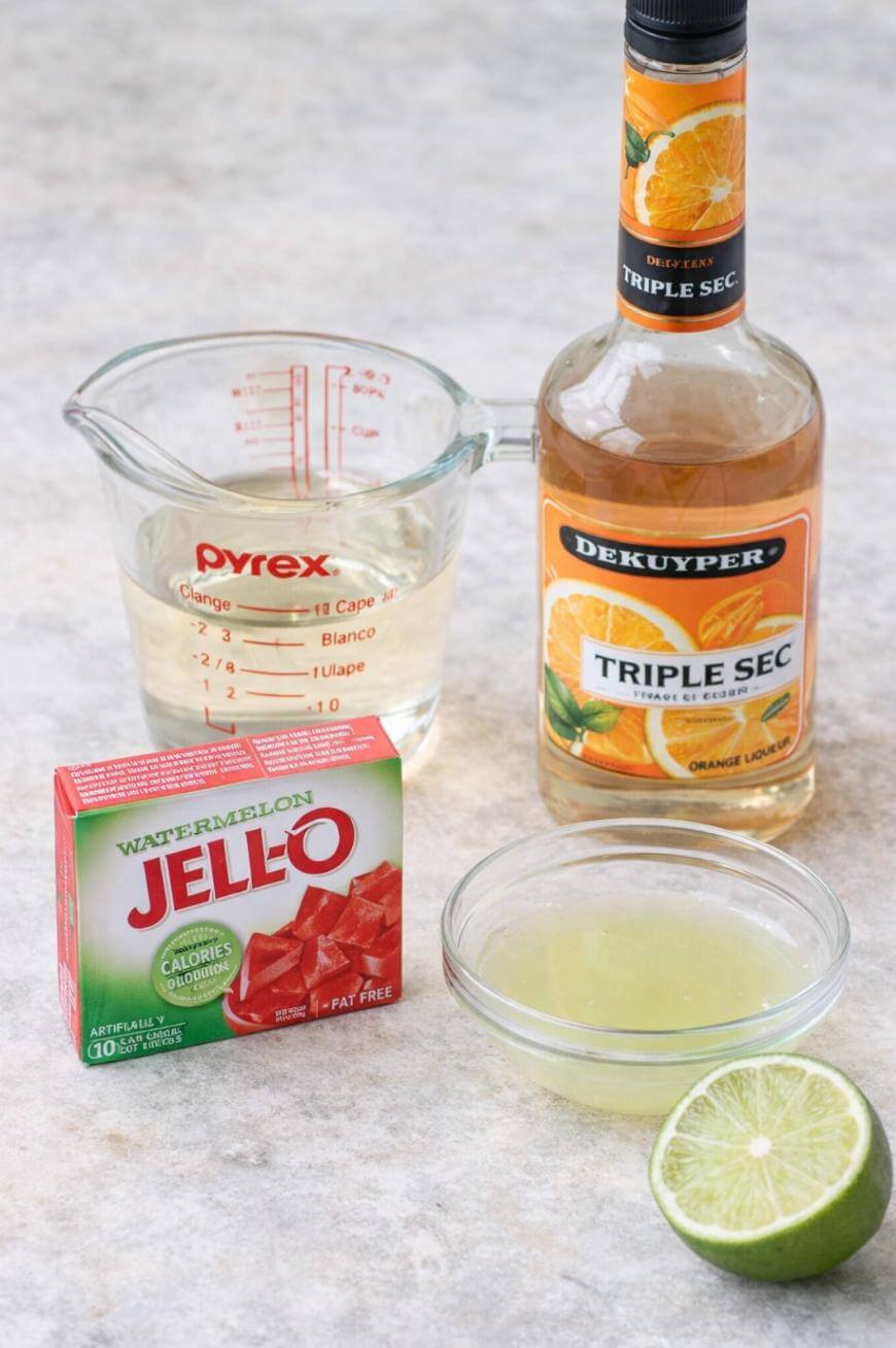 A bottle of triple sec, a halved lime, a bowl of lime juice, a measuring cup with clear liquid, and a box of watermelon Jell-O are arranged on a countertop.