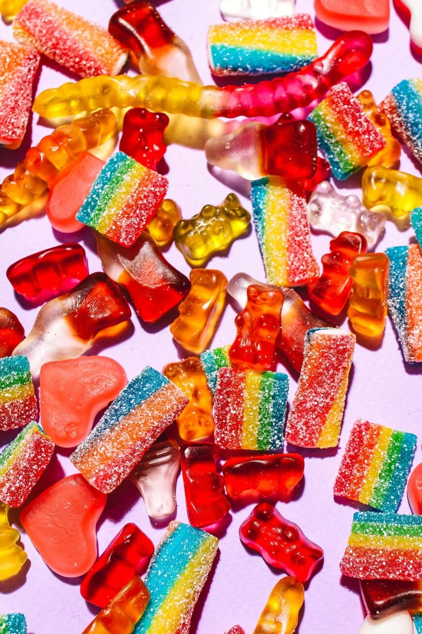 Assorted colorful gummy candies, including rainbow strips, gummy bears, gummy worms, and red heart-shaped gummies, scattered on a light purple surface.