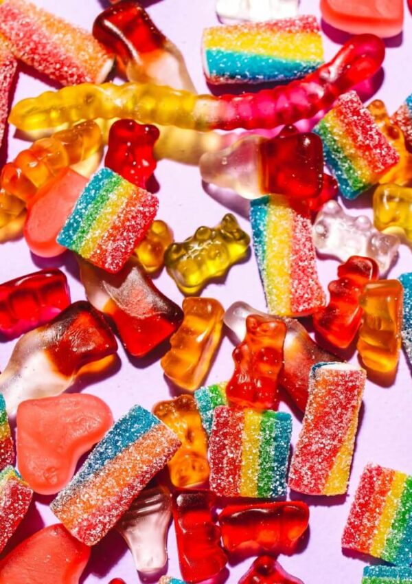 An assortment of colorful gummy candies, including gummy bears, rainbow-striped sour candies, a gummy worm, and heart-shaped gummies, scattered on a light purple background.