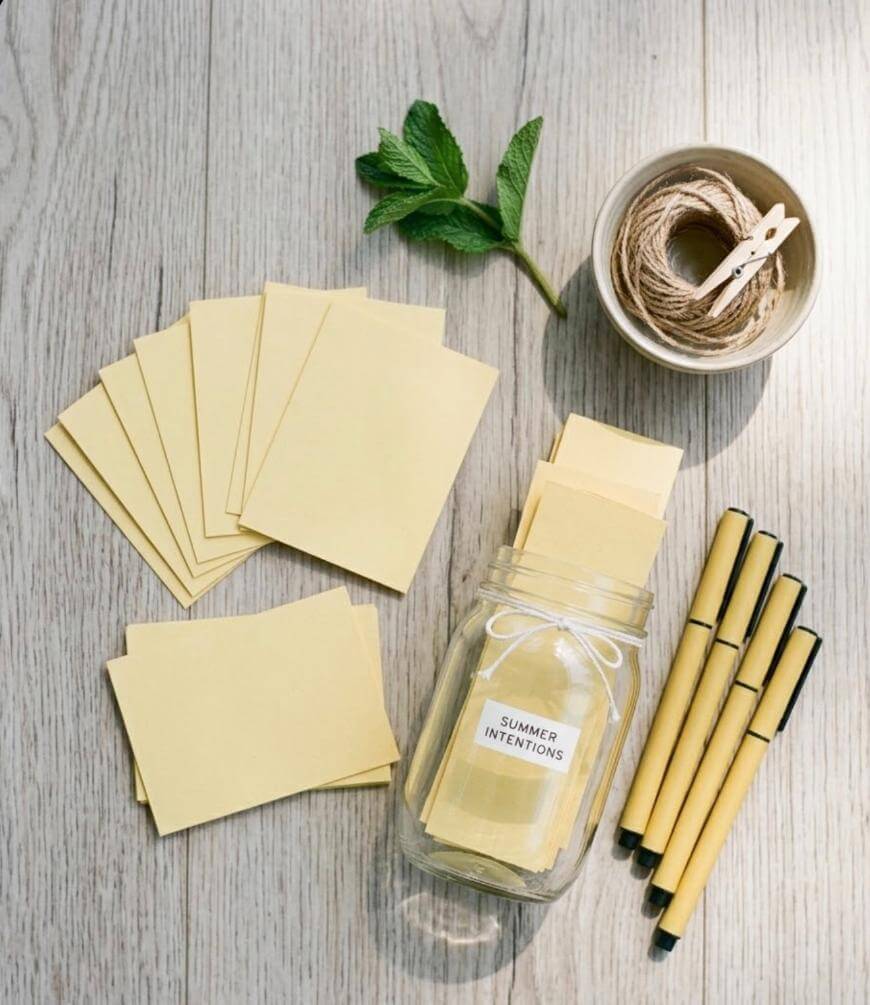 A jar labeled Summer Intentions is surrounded by blank yellow cards, four yellow pens, twine in a bowl with clothespins, and a mint sprig, all arranged on a light wooden surface.